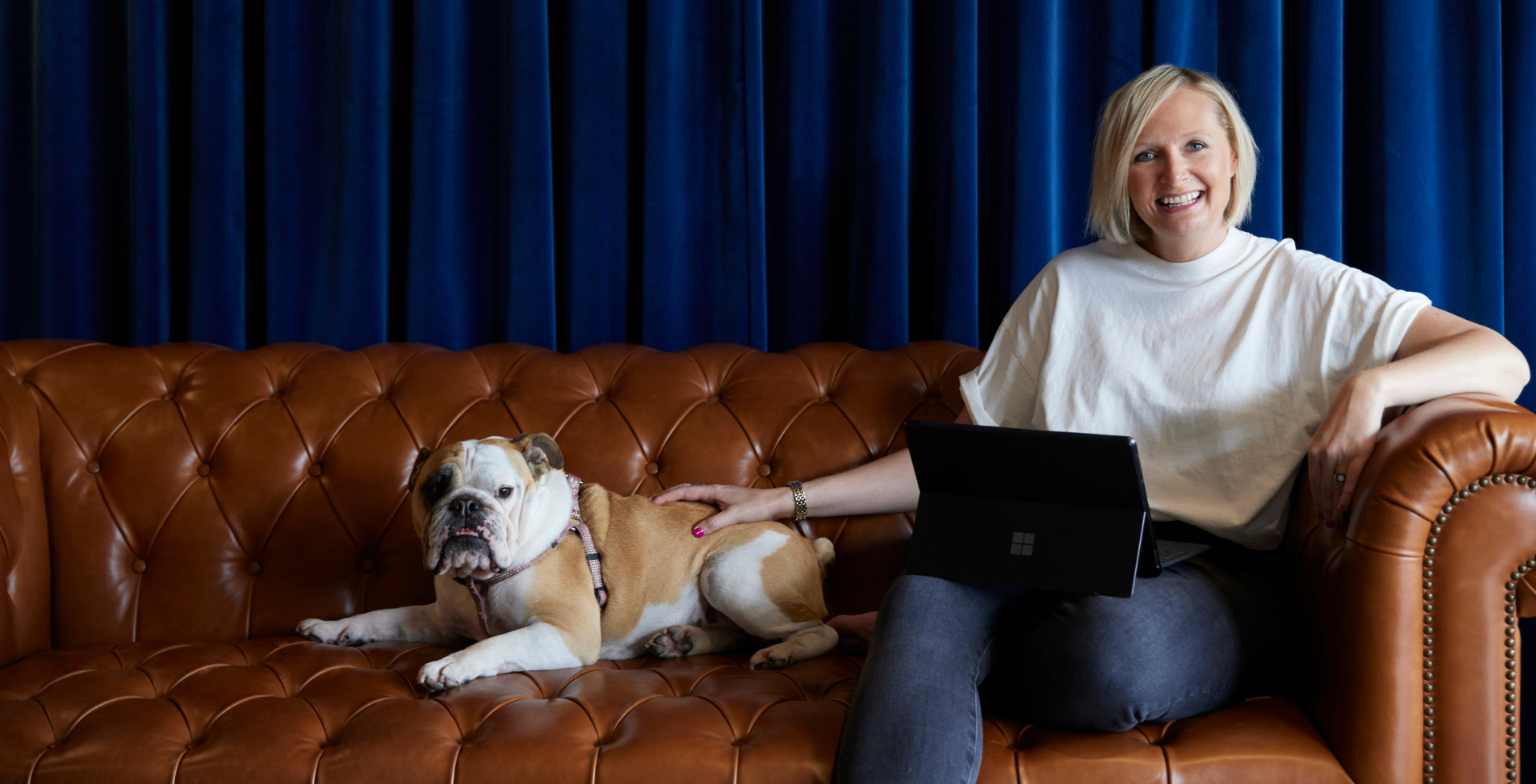 Woman sits on brown leather couch, petting a bulldog. She smiles, holding a tablet; blue wall backdrop.