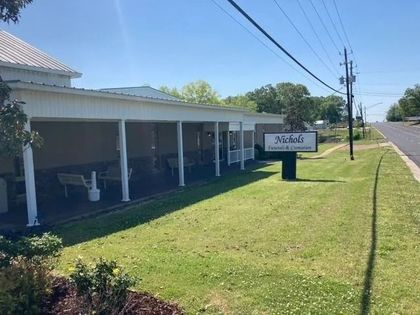 Single-story roadside building with white porch, green lawn, and a sign near the sidewalk under a clear blue sky