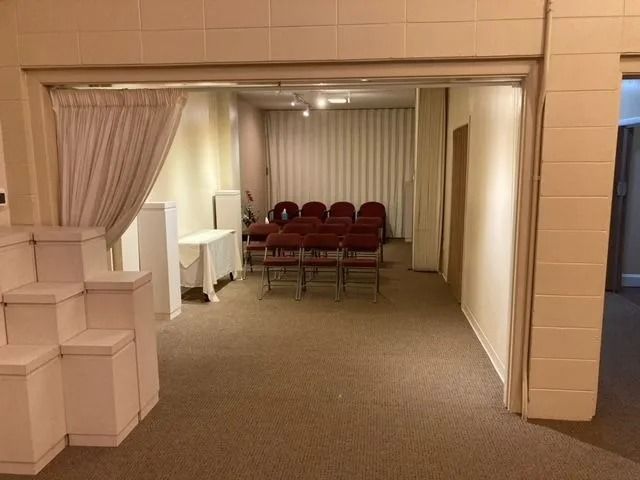 Small meeting room with red chairs, white curtains, and beige carpet, viewed through an open doorway