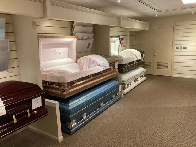 Rows of open caskets on display in a showroom, in maroon, white, and blue finishes