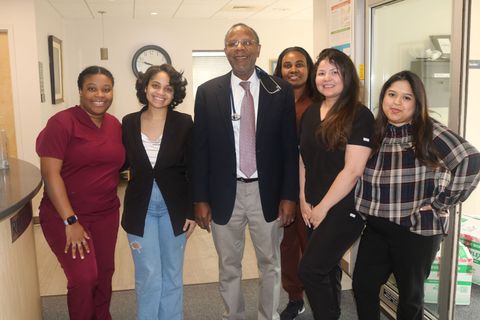 Group of six people posing in a medical office; includes a man in a suit, women in scrubs and casual attire.