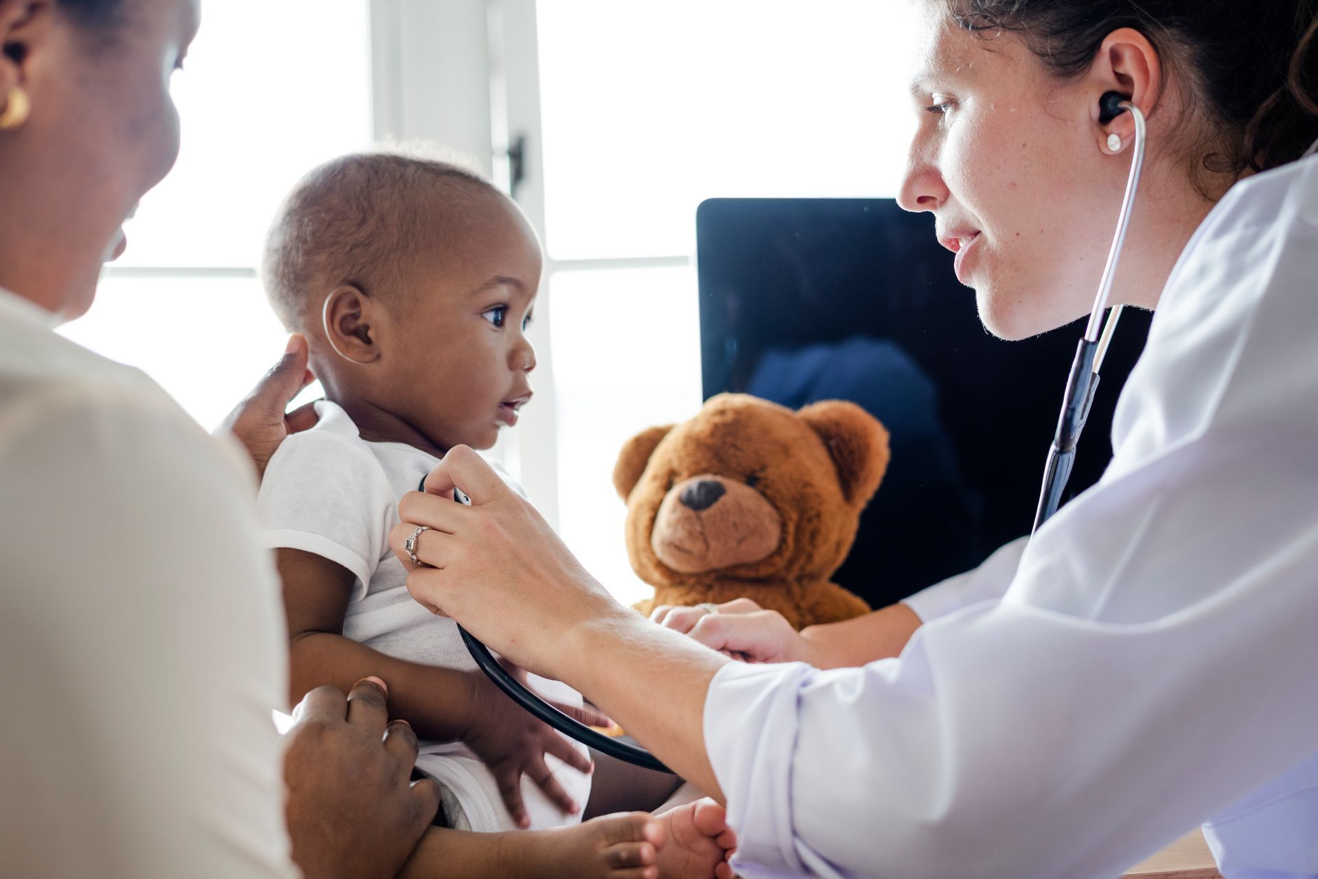 Doctor examining a baby while a parent holds the child, with a teddy bear nearby in a clinic room.