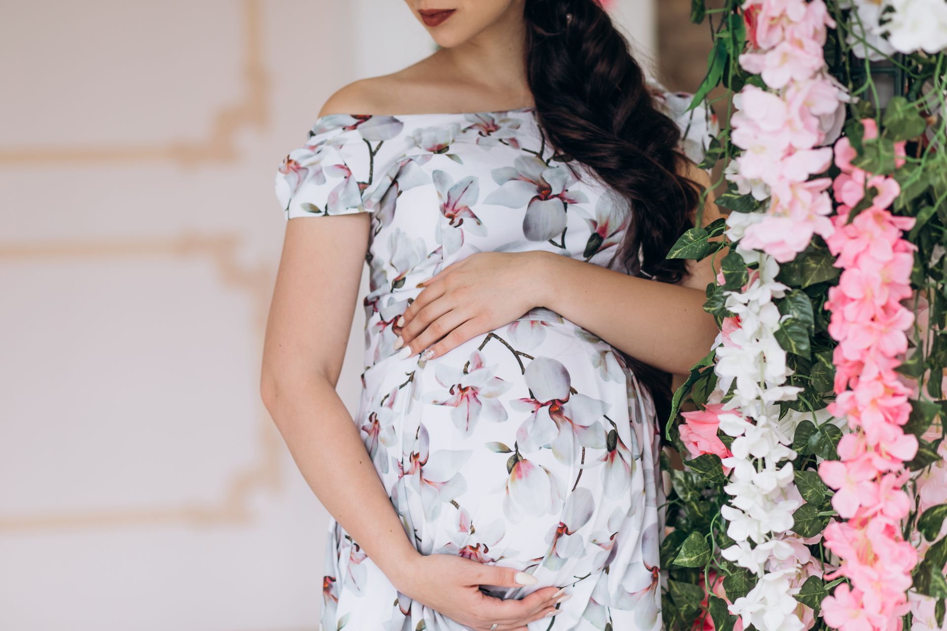 Pregnant woman in a floral dress posing beside pink flowers indoors