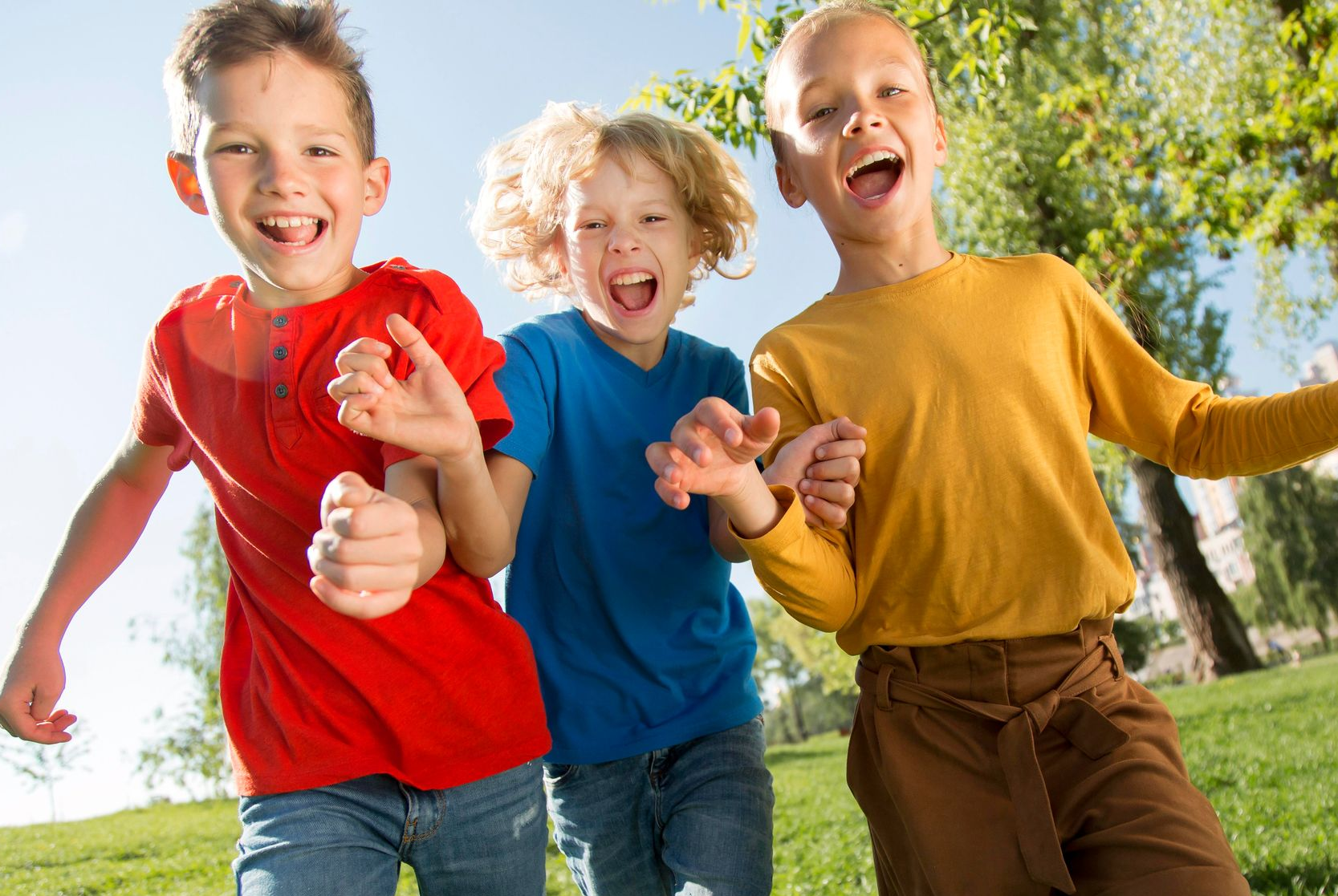 Three young boys in colorful shirts smiling and running through a park.