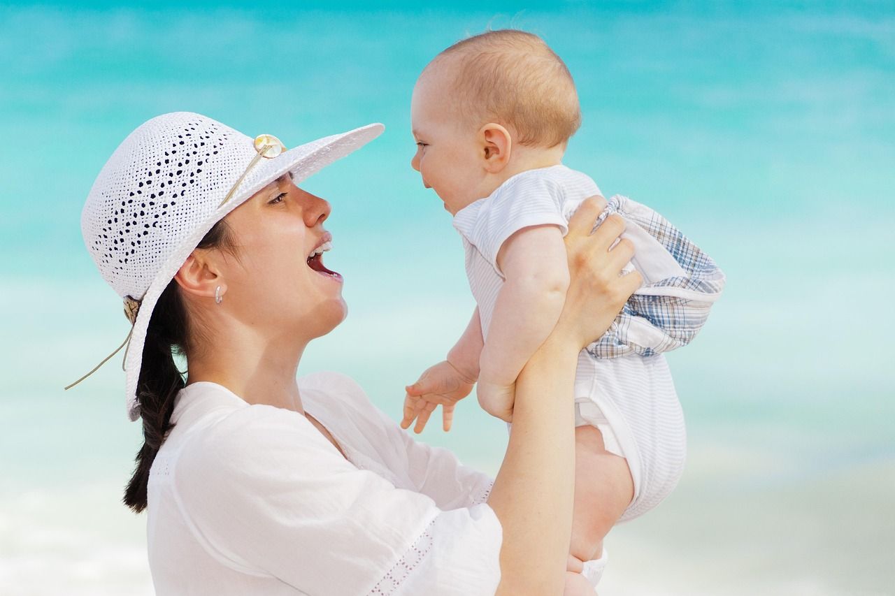Woman in a white hat holds up a baby on a beach. Both smile, blue water in the background.