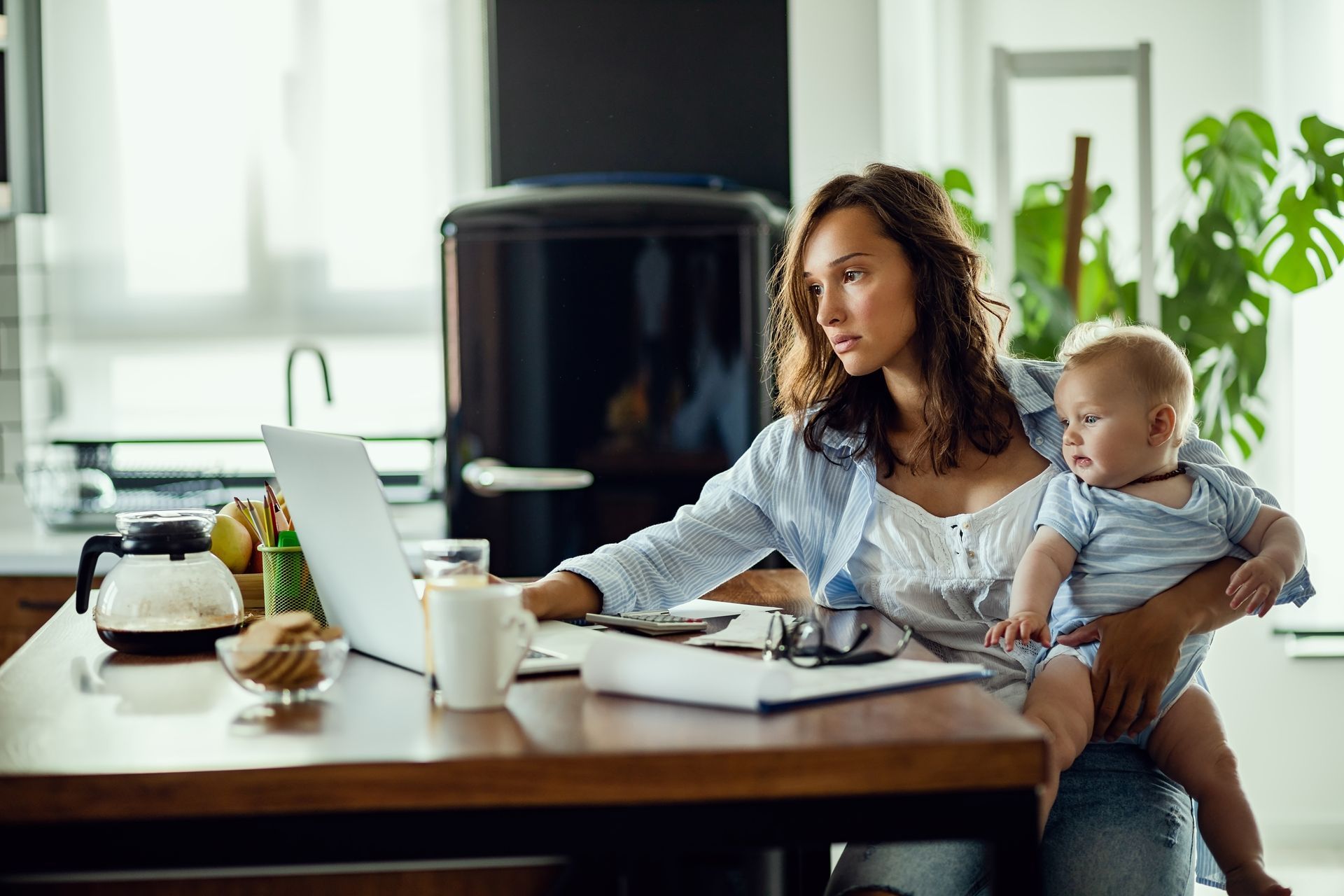 Woman working at a laptop while holding a baby at a kitchen table, with a coffee mug and notebook nearby.