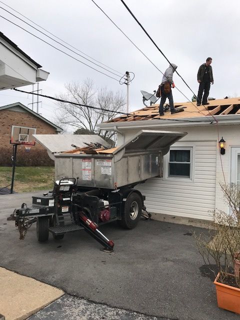 Workers on a house roof next to a roofing debris container. The container is extended from a trailer. Cloudy sky.