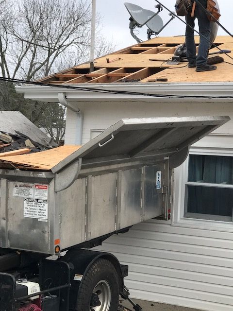Roofing work in progress: a worker on the roof, debris in a dumpster below, and a house.