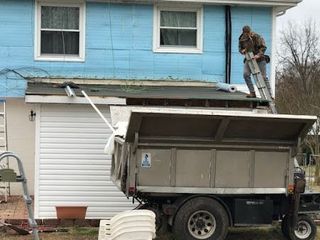 Man on ladder, removing roof. Dump truck beneath, blue house background.