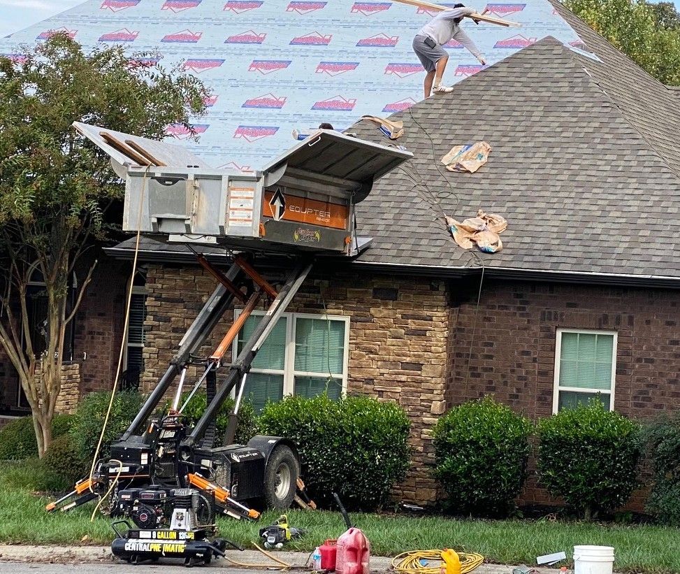 Worker installing asphalt shingles with a nail gun on a roof.