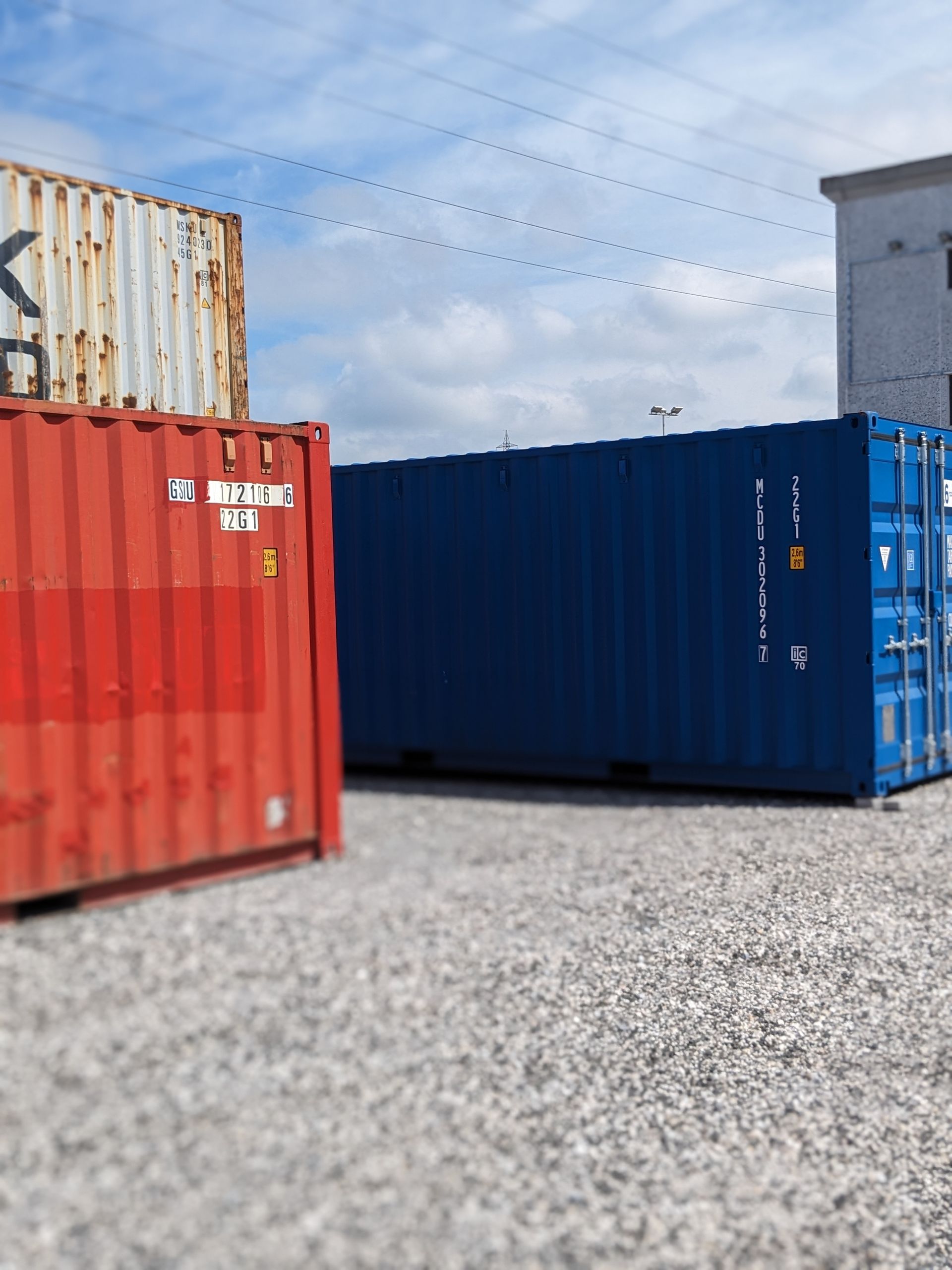 Red and blue shipping containers on gravel in an industrial yard under a partly cloudy sky