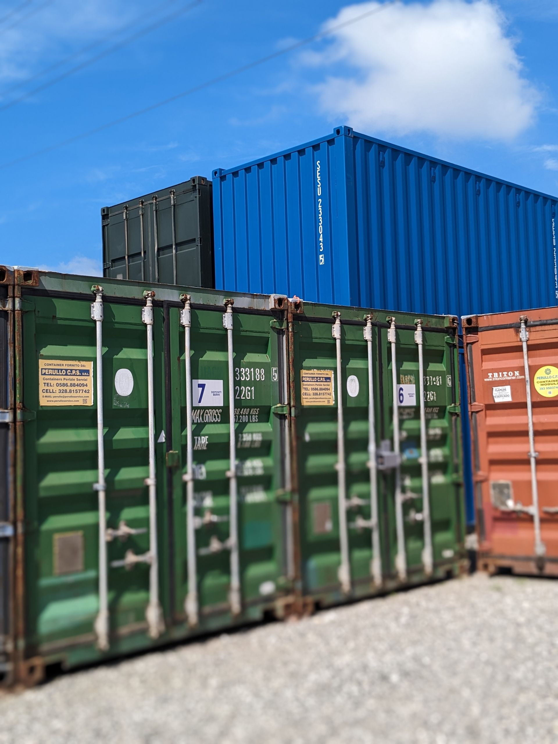 Rows of green, blue, and orange shipping containers in a gravel yard under a blue sky