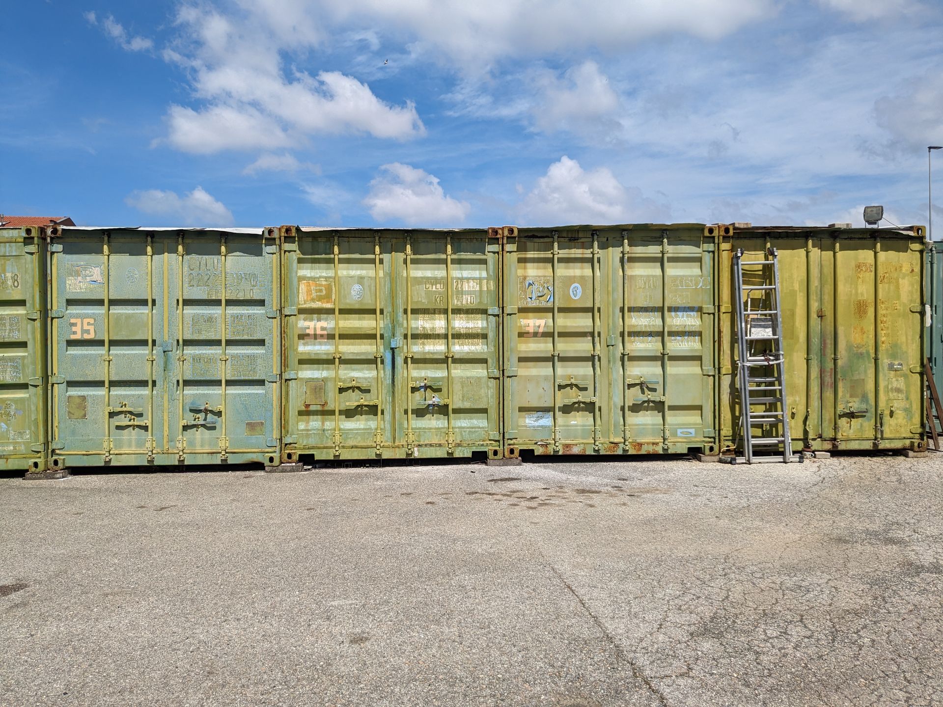 Weathered shipping containers in muted green and yellow on a gravel lot under a blue sky