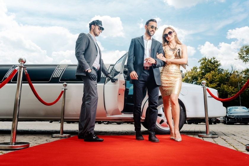 Three people posing on a red carpet beside a white limousine under a bright sky.