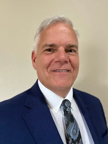 Man in a blue suit and patterned tie smiling against a beige background