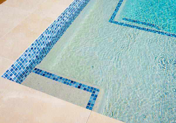 Blue mosaic-lined swimming pool corner with clear rippling water and beige deck tiles