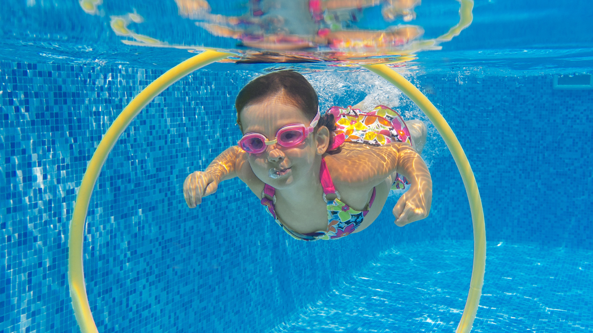 Child swimming through a yellow hoop underwater in a blue pool, wearing pink goggles and a colorful swimsuit