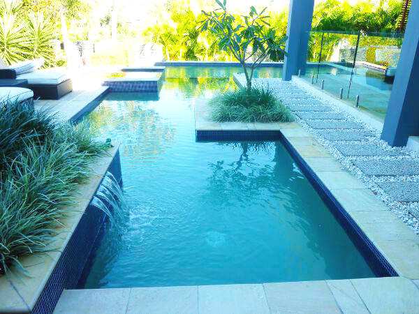 Modern backyard pool with turquoise water, lounge chairs, and lush greenery in bright sunlight