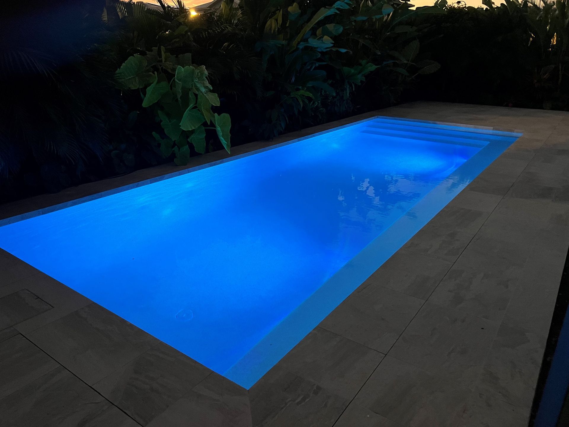 Rectangular blue-lit swimming pool at night, surrounded by dark patio tiles and greenery