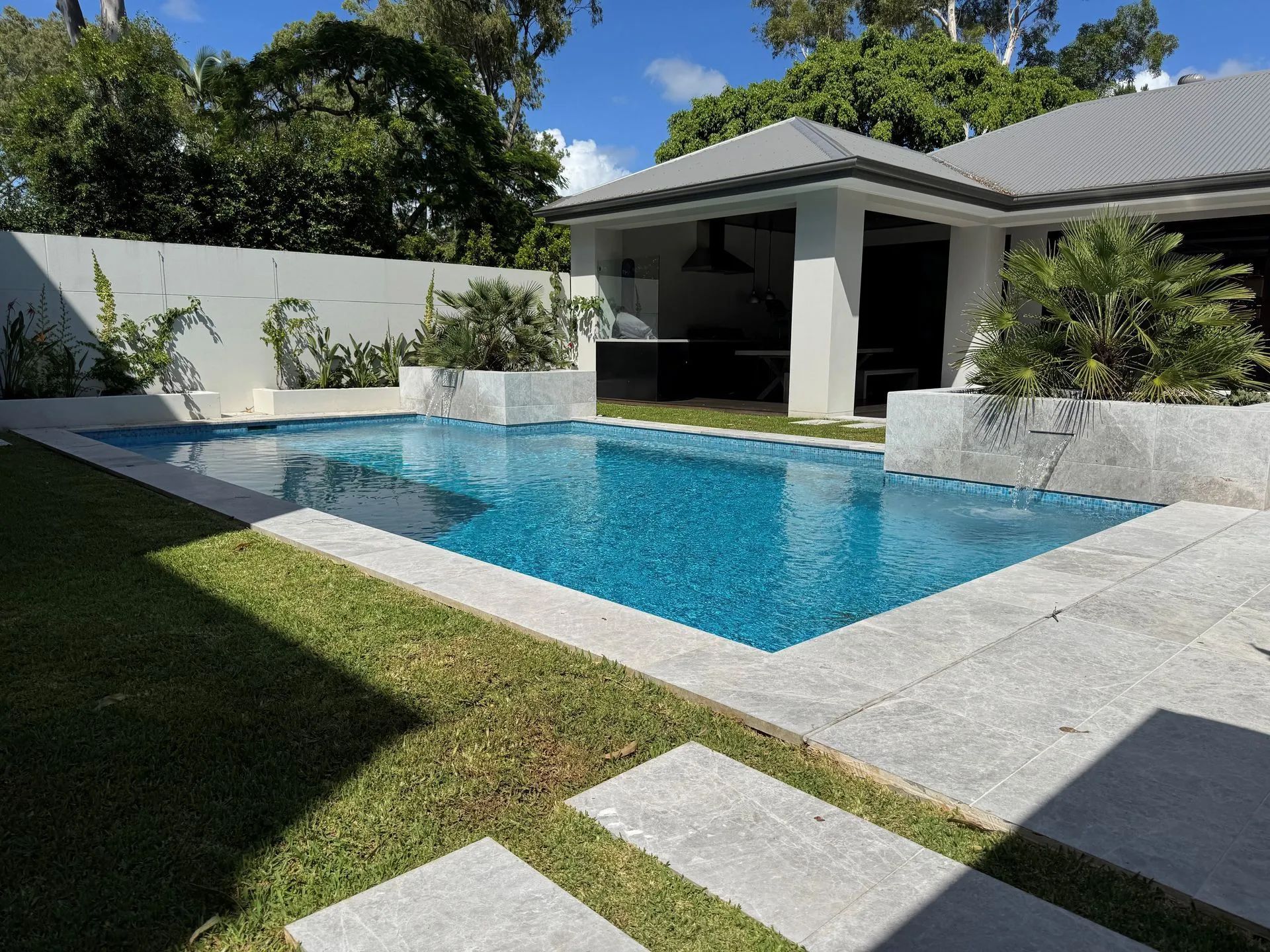 Backyard with a rectangular blue swimming pool, white patio, and shaded modern poolside pavilion.