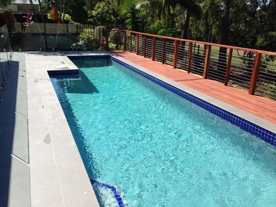 Long narrow outdoor pool with blue water, white tile deck, and wooden railing beside lush greenery.