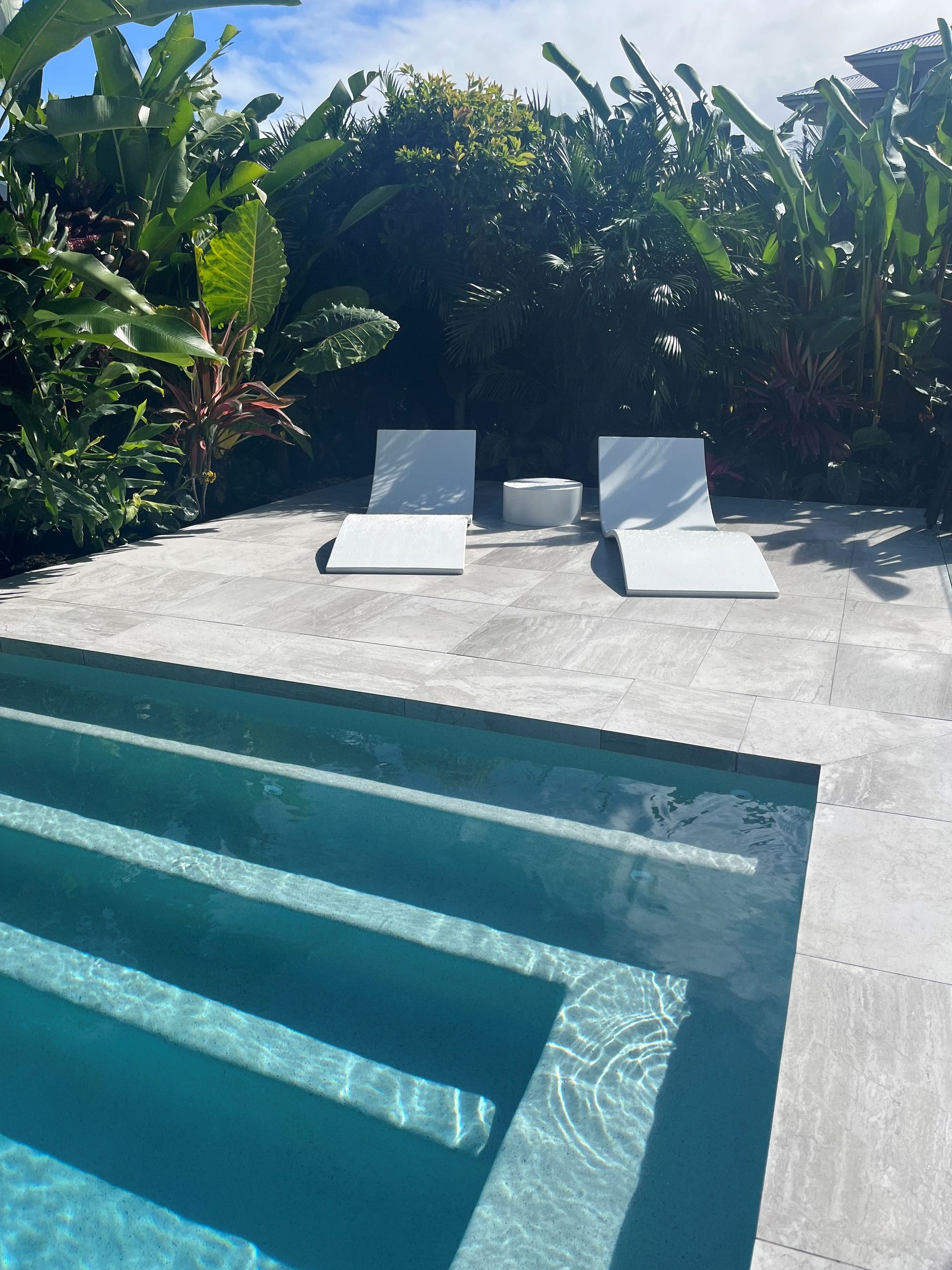 Modern pool deck with two white lounge chairs beside lush tropical greenery