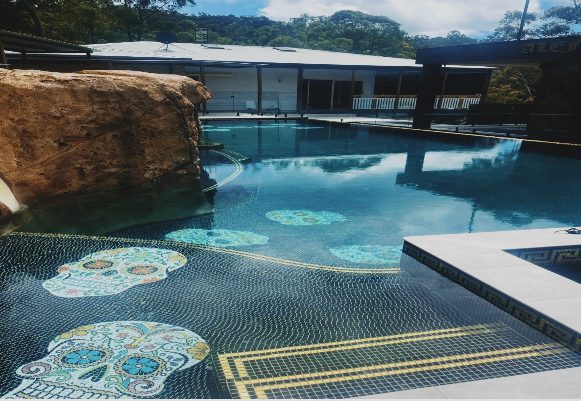 Outdoor pool with clear blue water, rock waterfall, and decorative mosaic tiles under a covered pavilion.