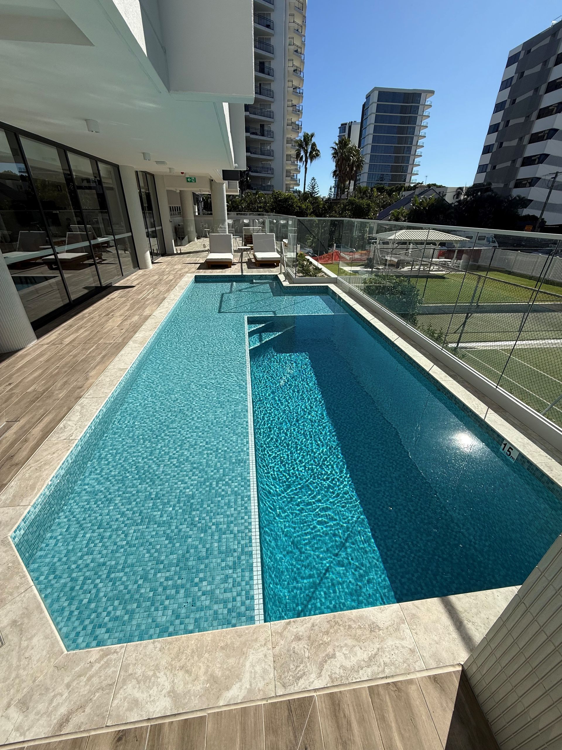 Outdoor swimming pool with turquoise water beside modern apartment buildings and lounge chairs on a sunny terrace