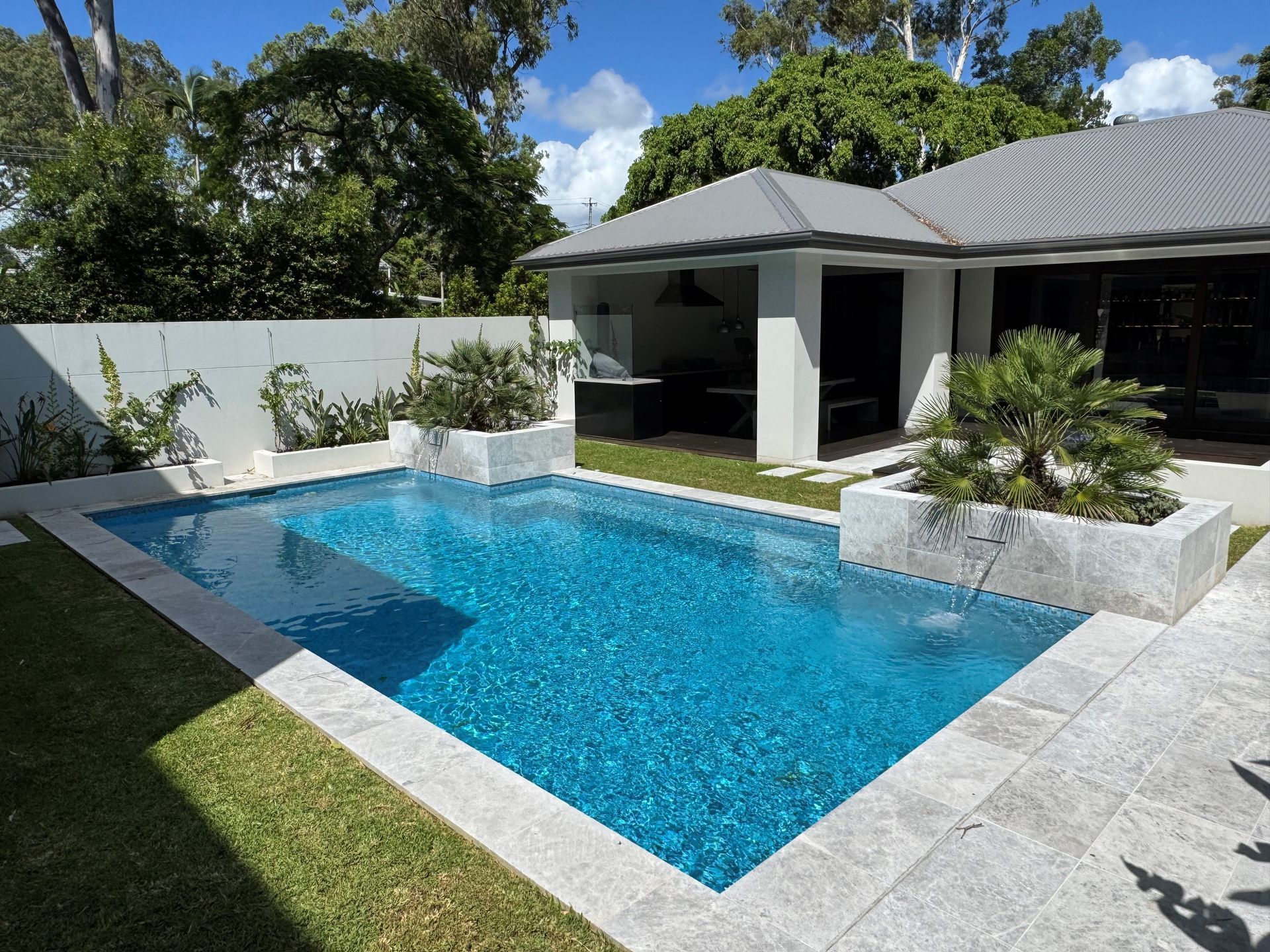 Modern backyard with a rectangular blue pool, white stone patio, and covered outdoor lounge area.