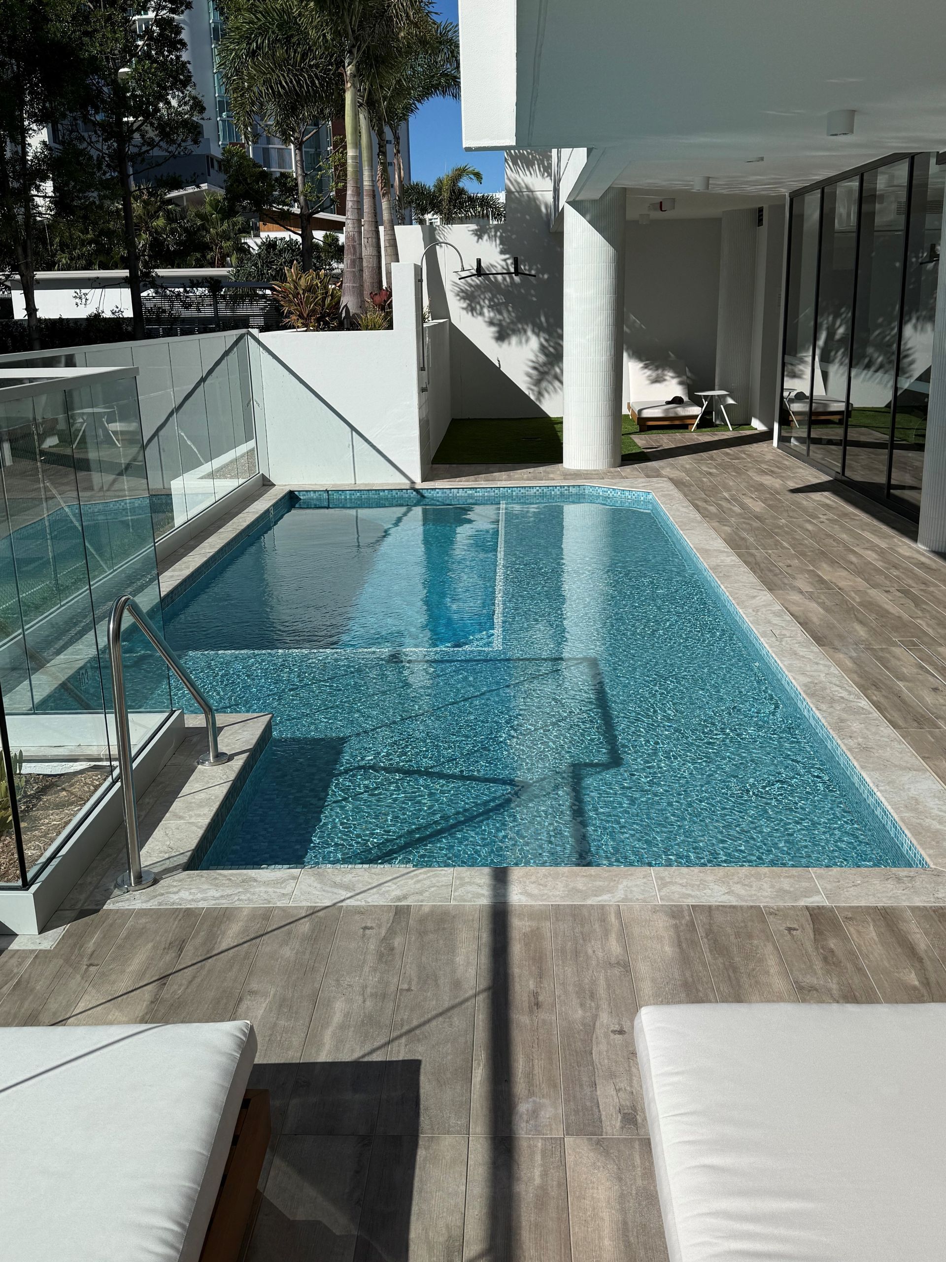 Modern backyard pool with clear blue water, glass railing, and white lounge chairs in bright sunlight