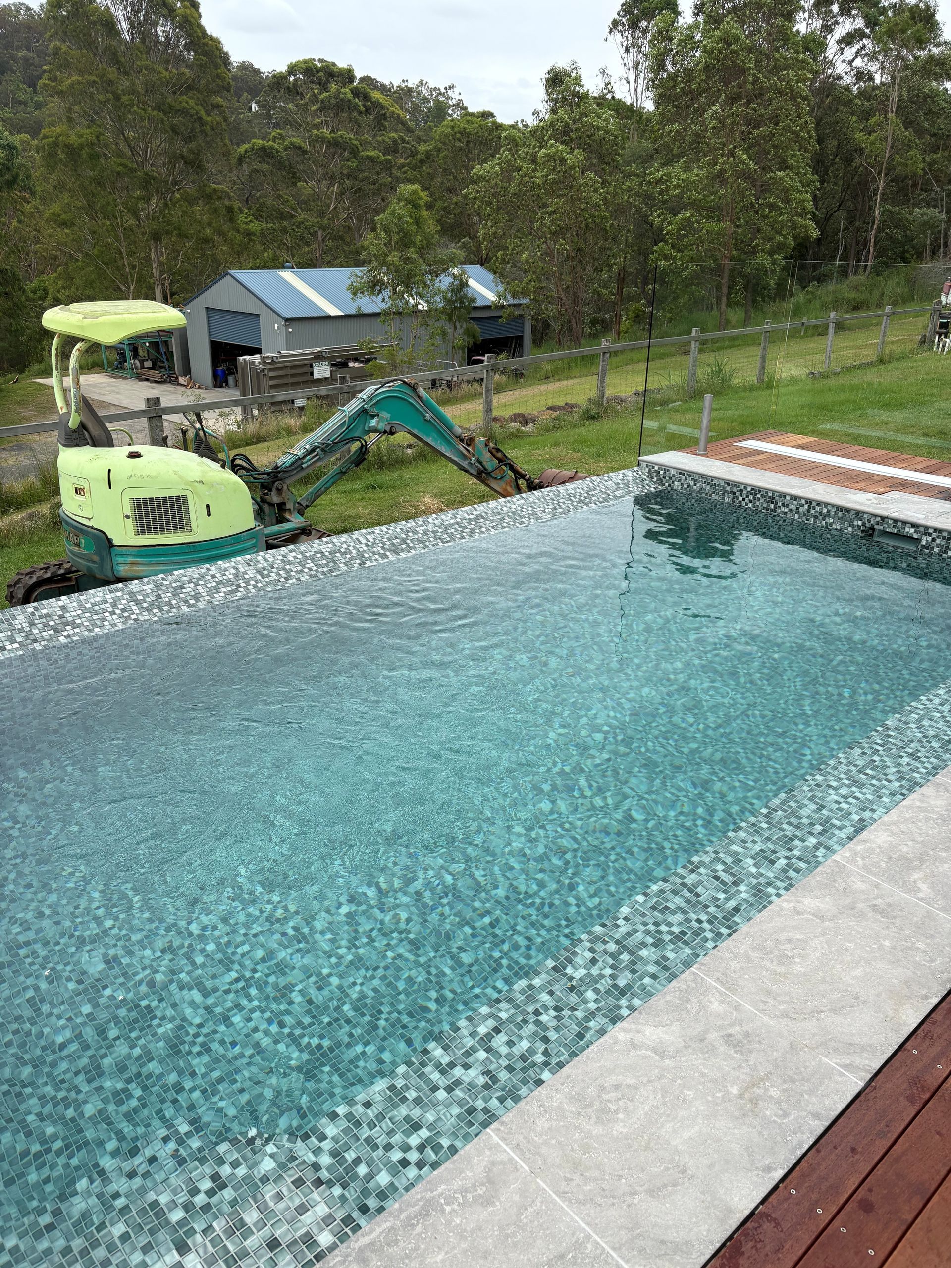 Backyard pool with turquoise water, a green excavator nearby, and a small shed in the background