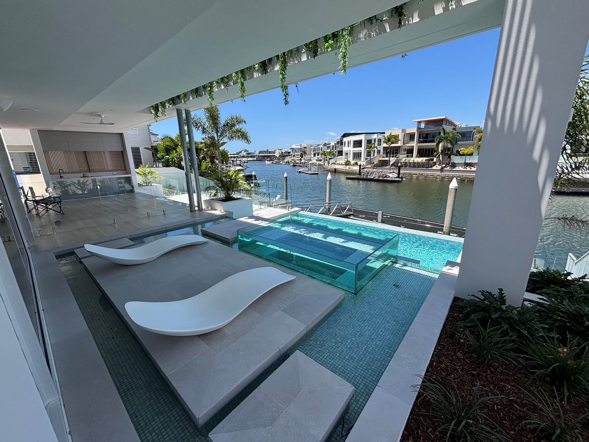 Modern waterfront patio with two white lounge chairs and a turquoise infinity pool overlooking a canal