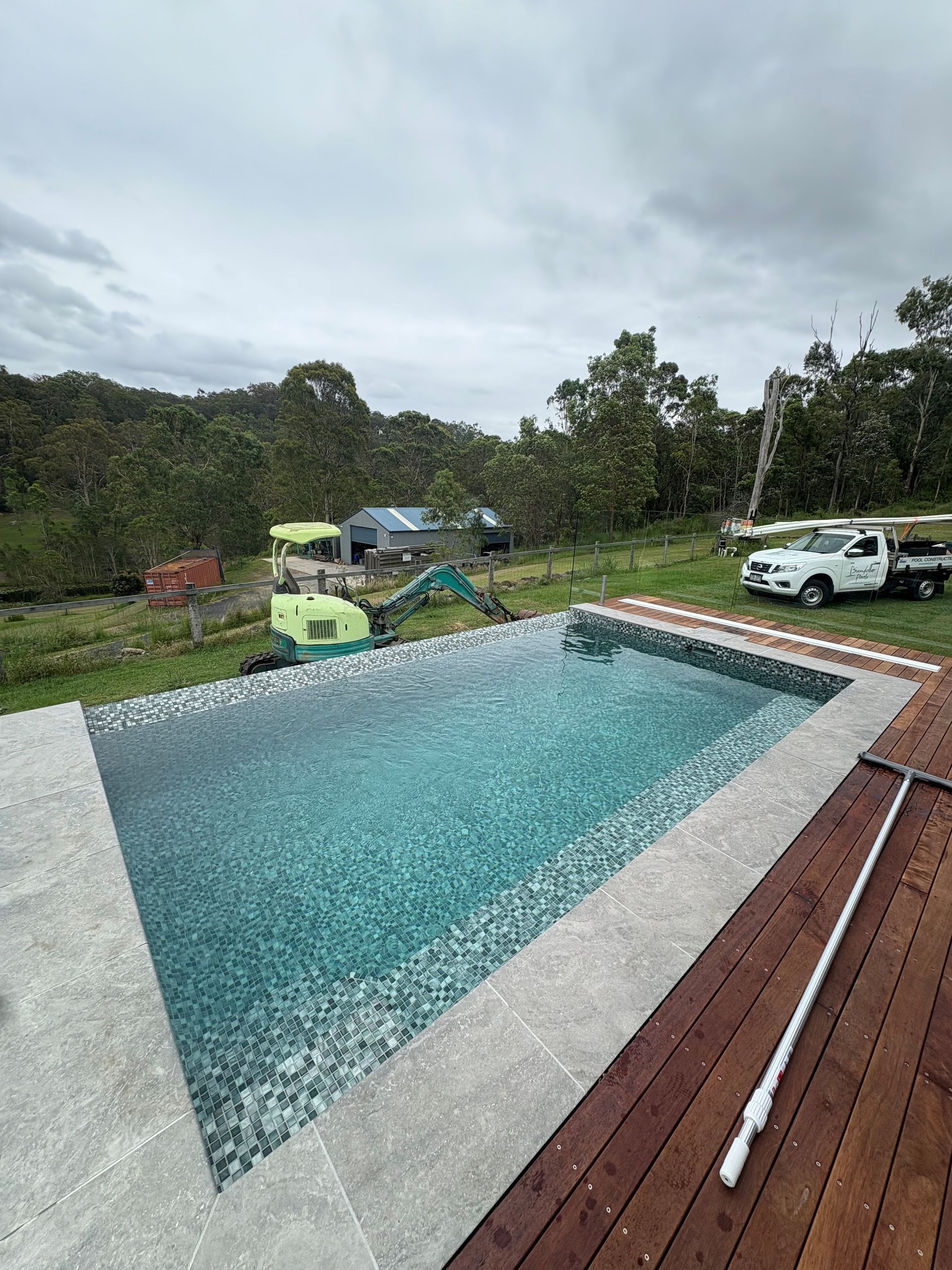 Backyard pool with blue water, white deck, trucks, and trees under an overcast sky