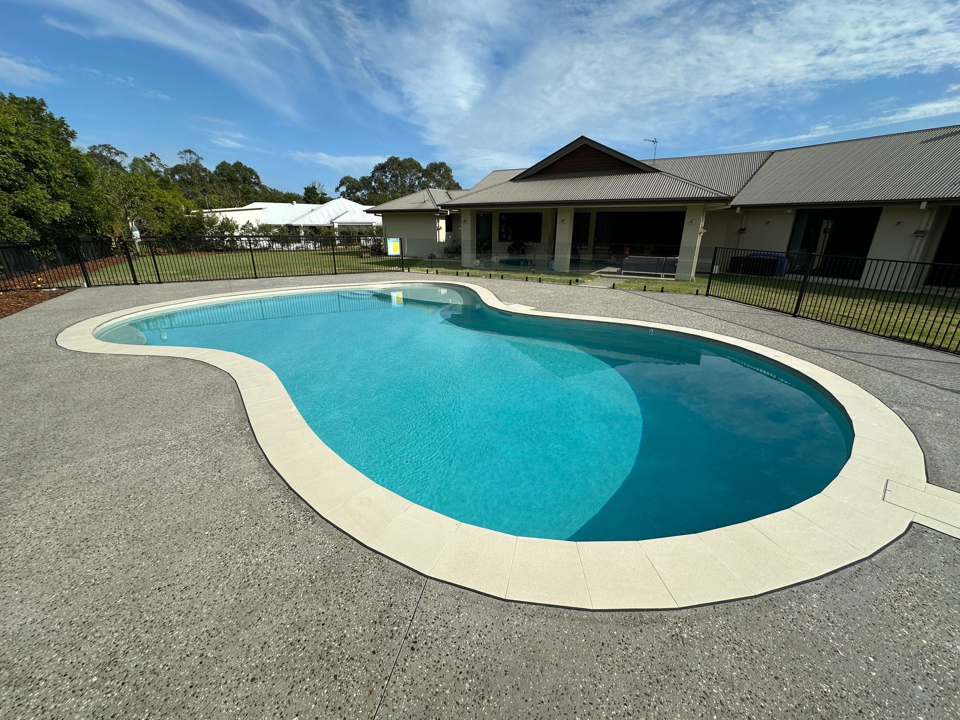 Kidney-shaped outdoor swimming pool beside a building and trees under a clear sky