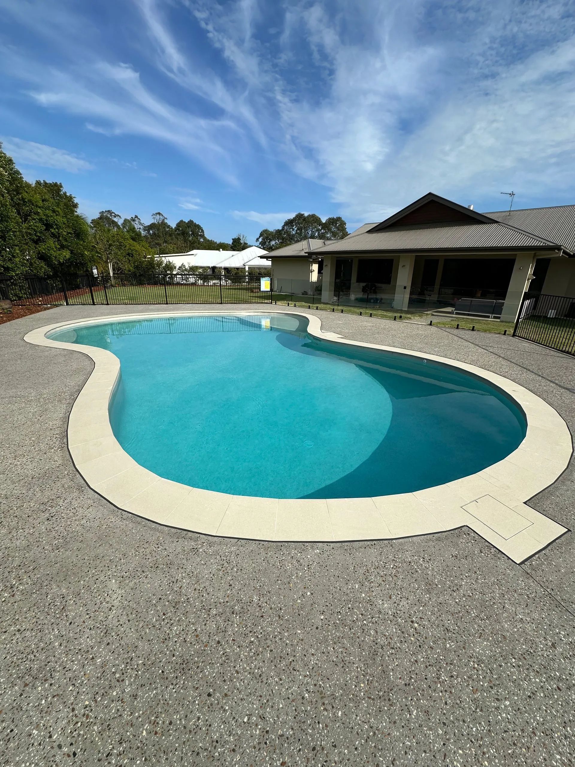 Curved turquoise backyard pool with white coping, gravel deck, and a patio under a blue sky