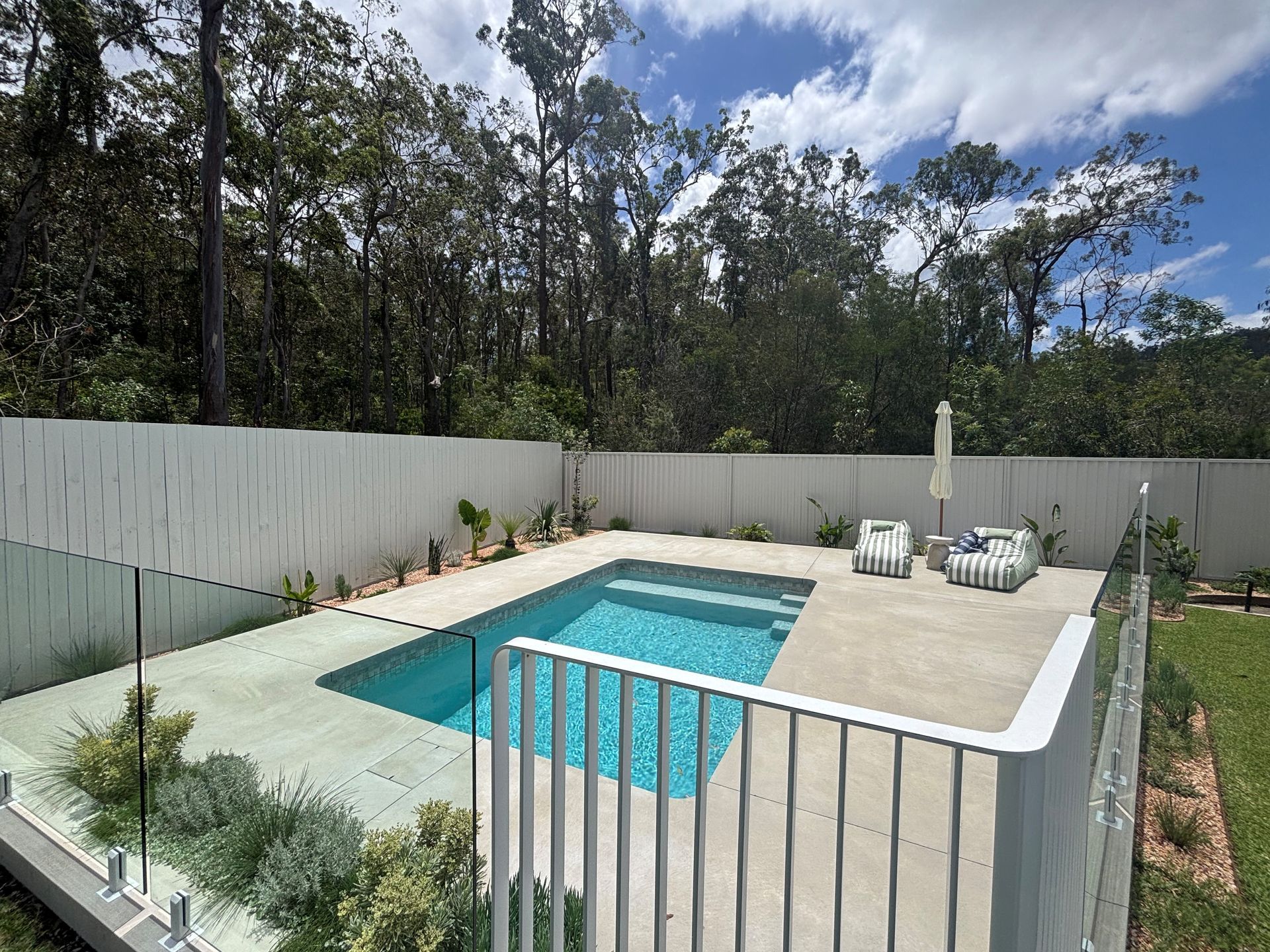 Backyard pool with white patio, lounge chairs, and tall fenced privacy wall under a cloudy sky
