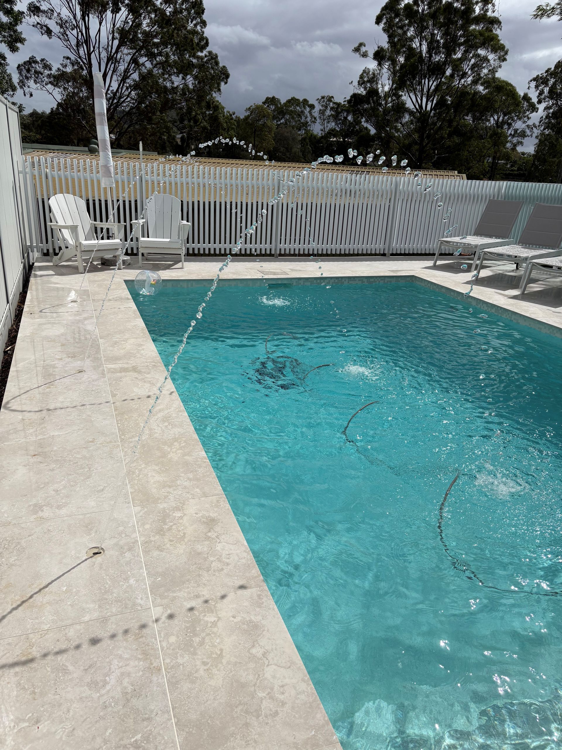 Poolside patio with turquoise pool water, white fence, lounge chairs, and trees in the background