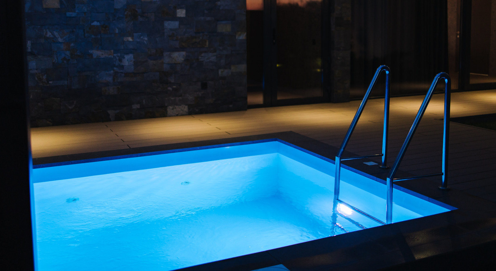 Blue-lit indoor swimming pool with metal handrails beside a dark wall at night
