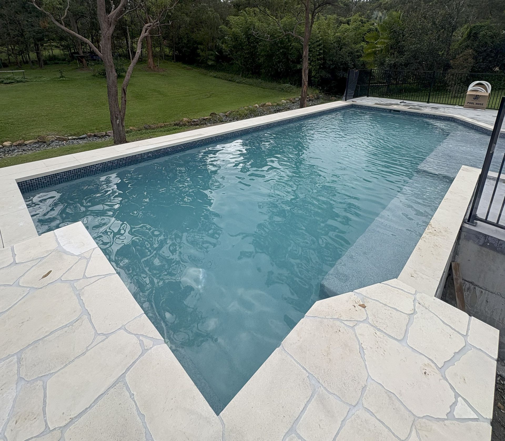 Backyard swimming pool with blue water and white stone decking beside a grassy yard.