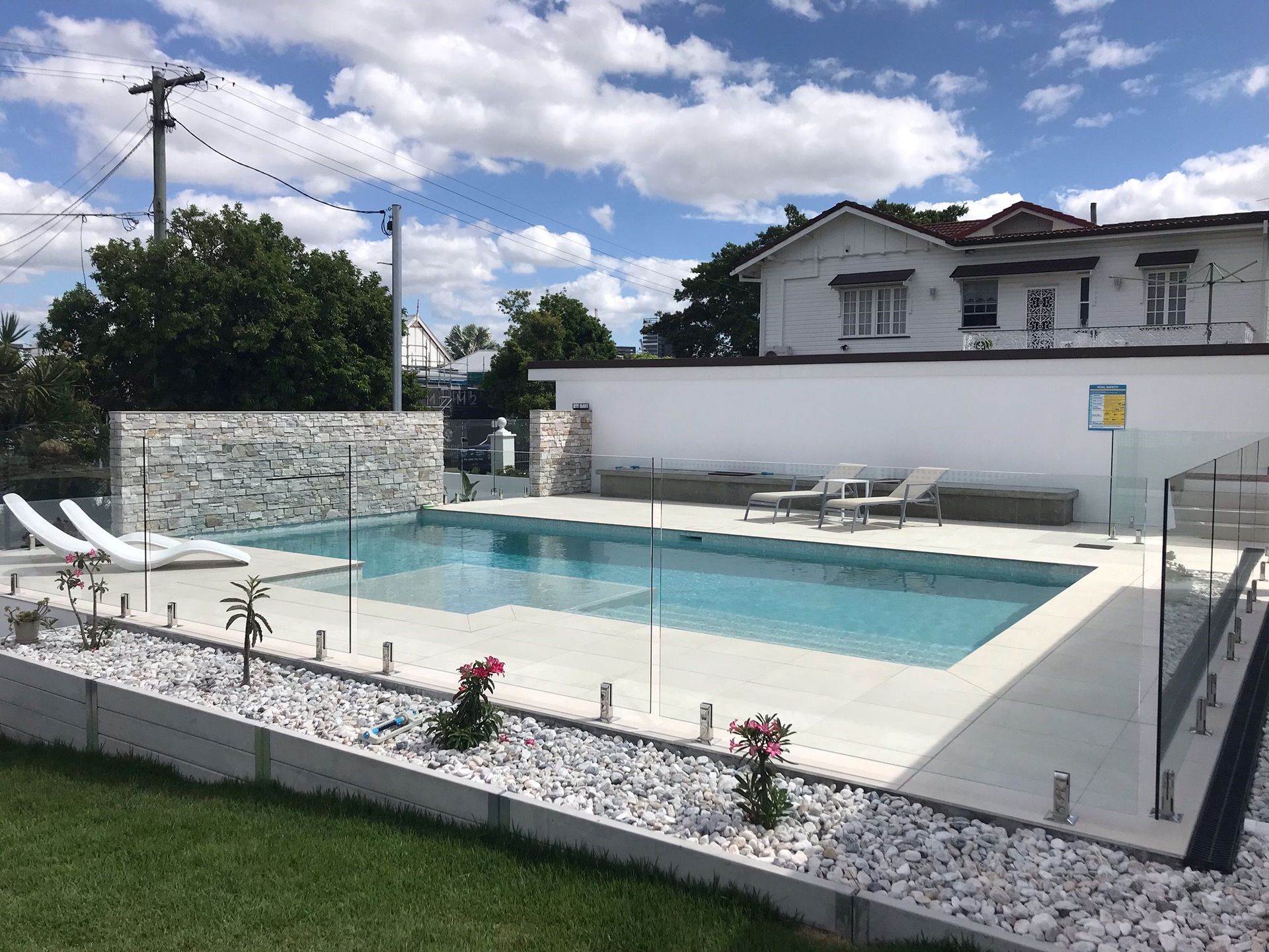 Backyard pool with lounge chairs, white patio walls, and a house under a partly cloudy sky