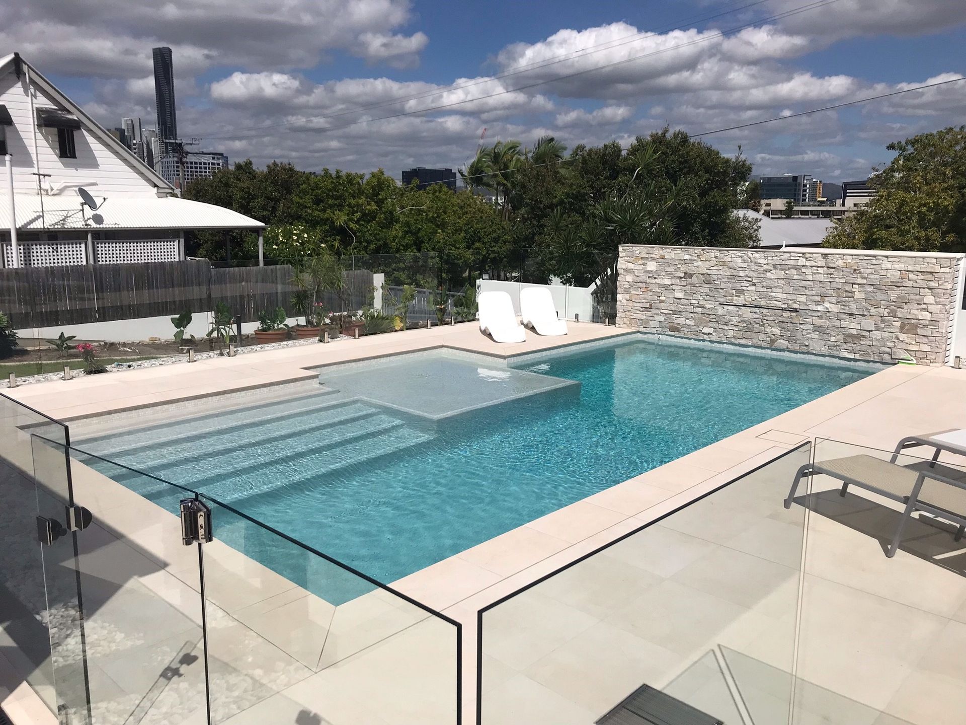 Modern backyard pool with glass railing, white loungers, and stone wall under a cloudy sky