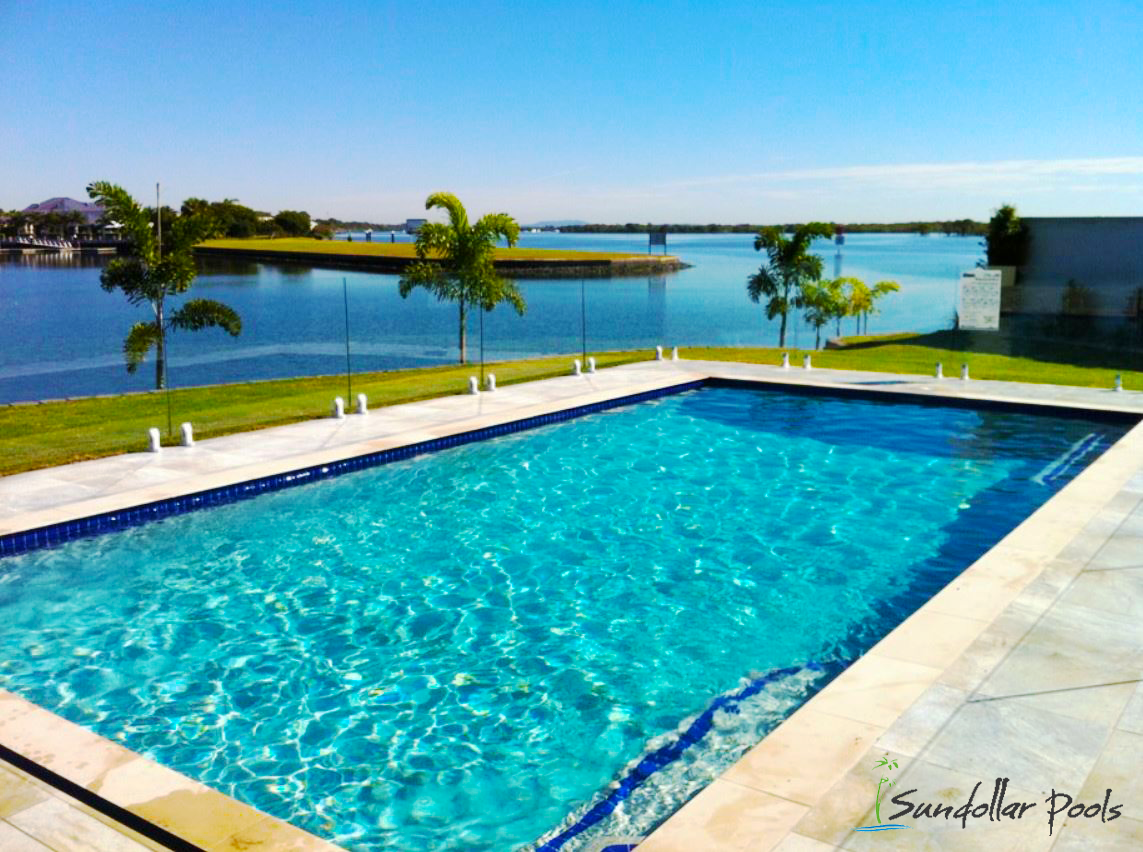 Blue swimming pool beside a calm bay, with palm trees and a bright sky beyond a white deck.