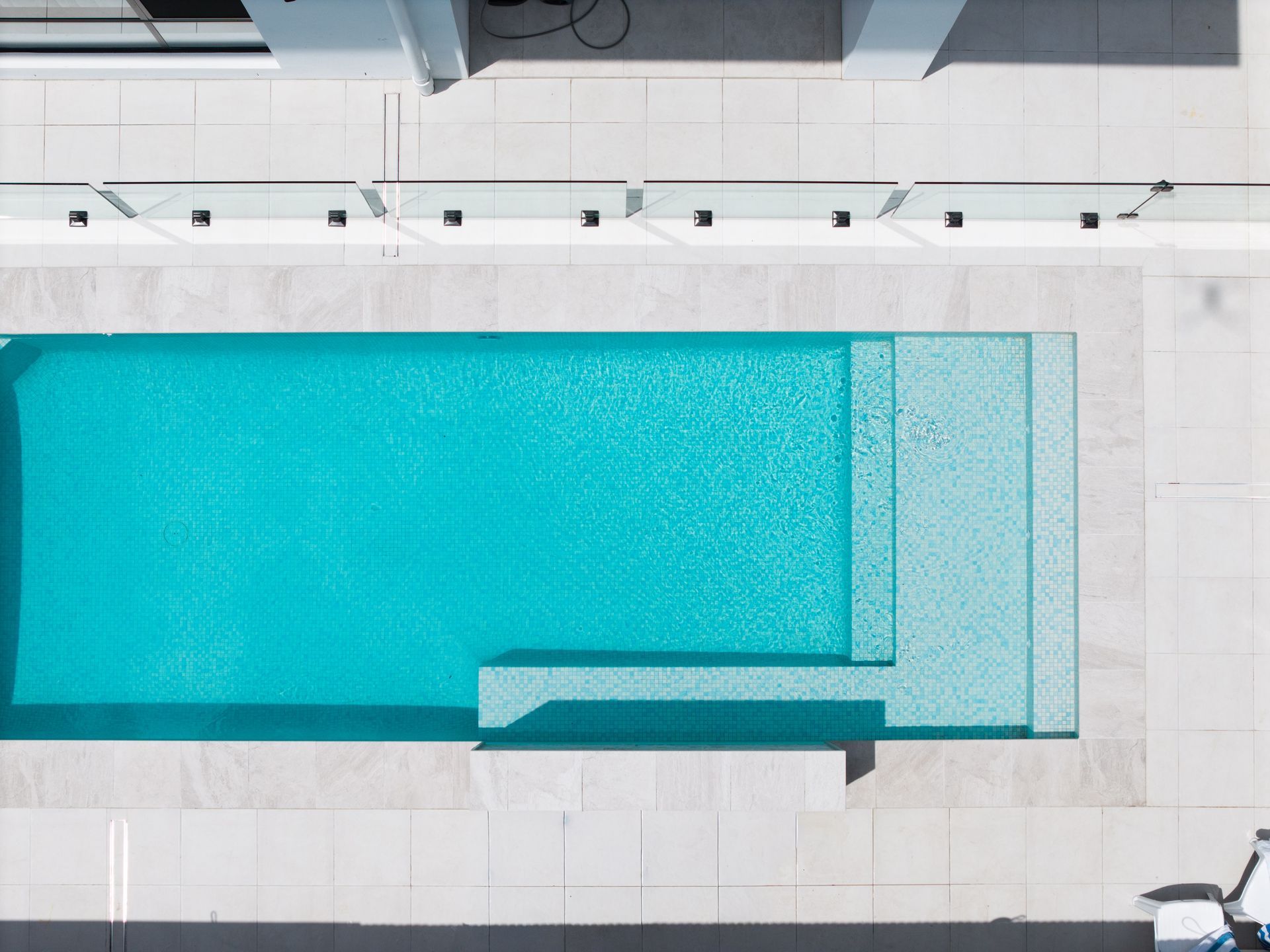Top-down view of a turquoise swimming pool with a shallow section and white tiled deck.