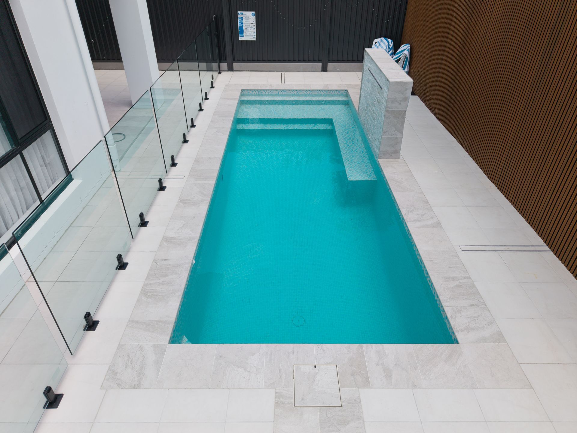 Narrow turquoise pool on a white tiled rooftop terrace with glass railing and brown privacy wall
