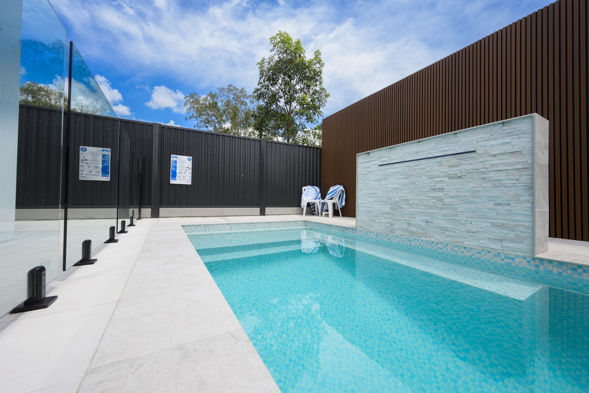 Outdoor pool with turquoise water, white deck, dark wood fence, and two white lounge chairs under a blue sky