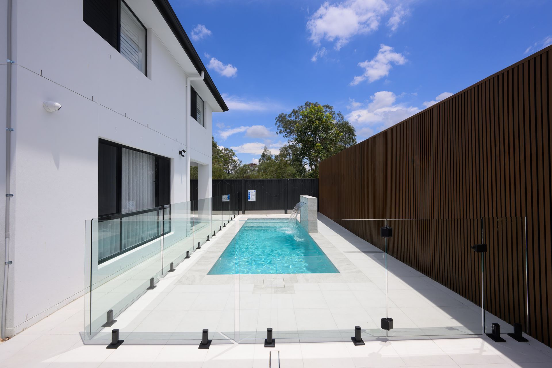 Modern house courtyard with a turquoise swimming pool, glass railing, and tall wooden fence under a blue sky