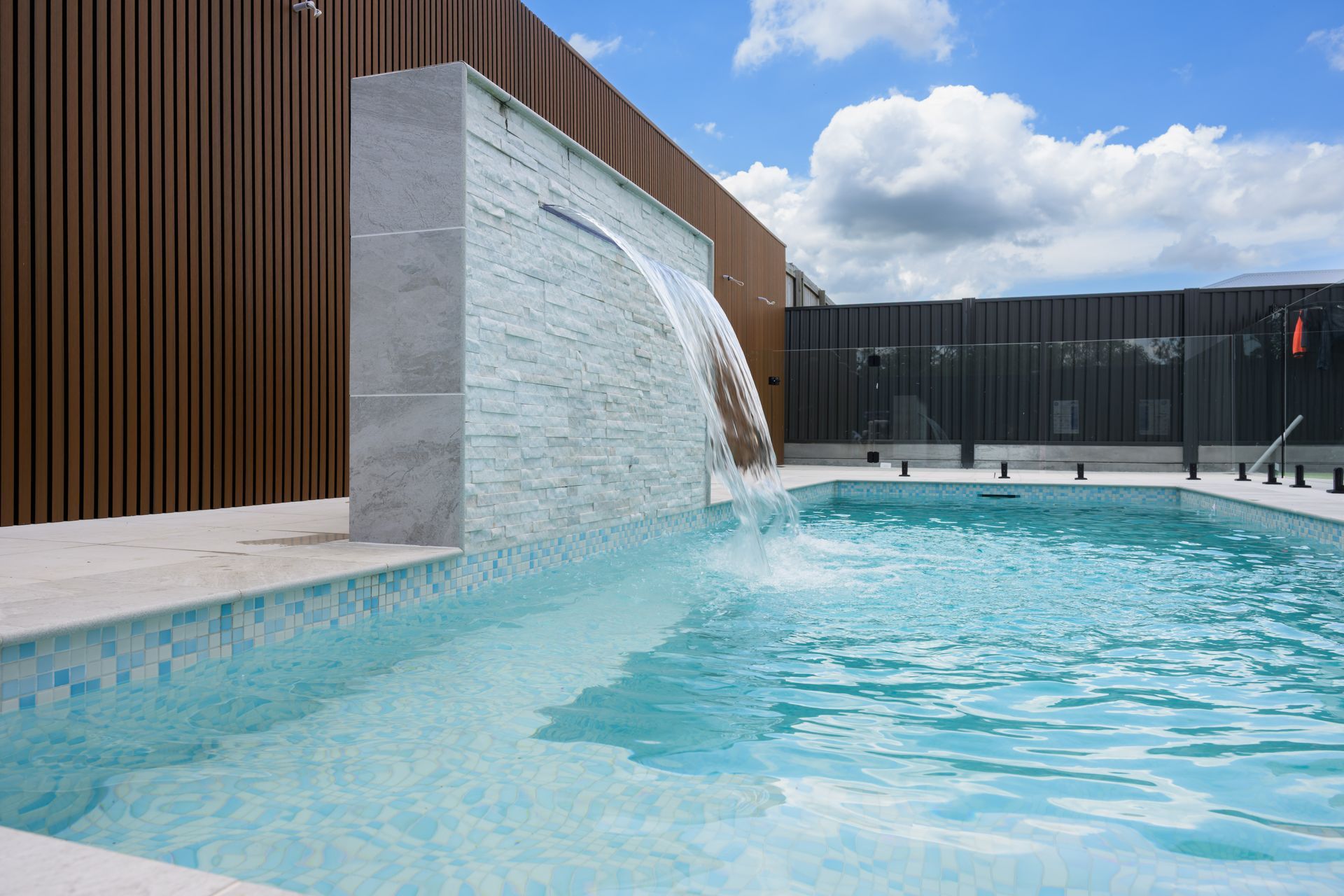 Modern outdoor pool with a stone waterfall and blue water beside a wooden wall