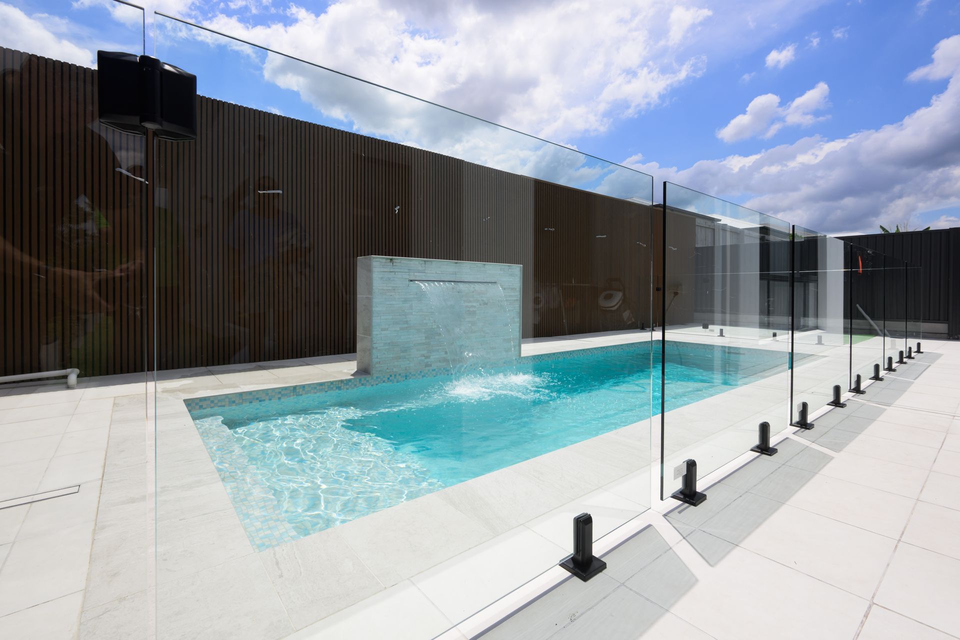 Modern rooftop pool with turquoise water, glass safety fence, and dark wood privacy wall under a cloudy sky