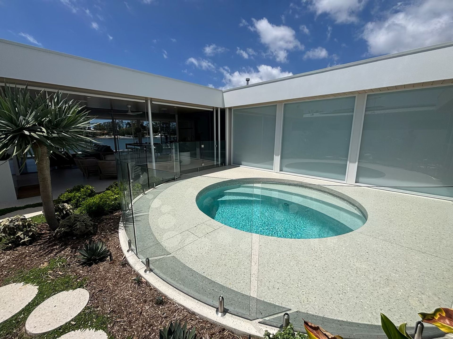 Modern patio with a small circular turquoise pool, glass railing, and white privacy walls under a blue sky