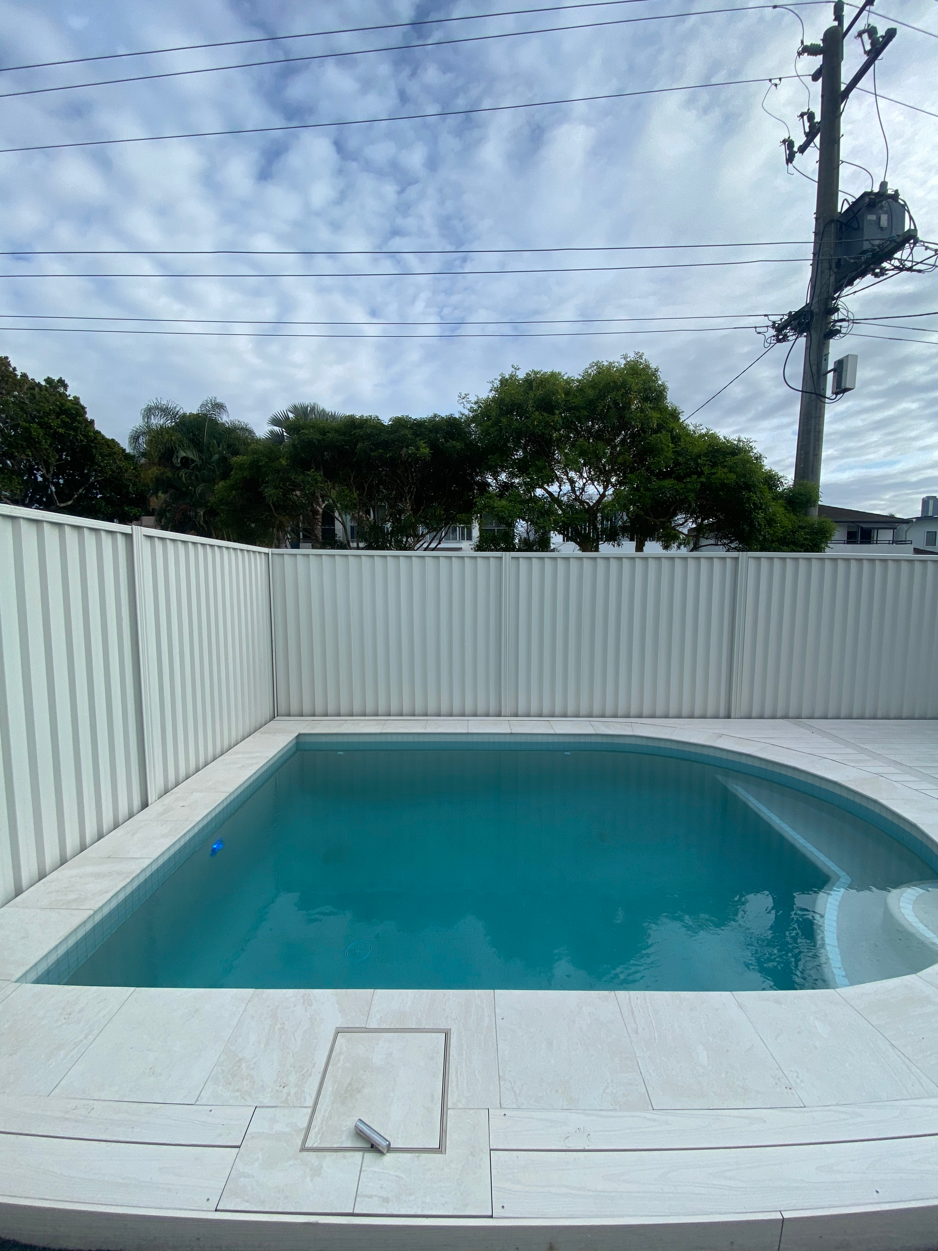 Small backyard swimming pool with turquoise water, white fence, and cloudy sky overhead